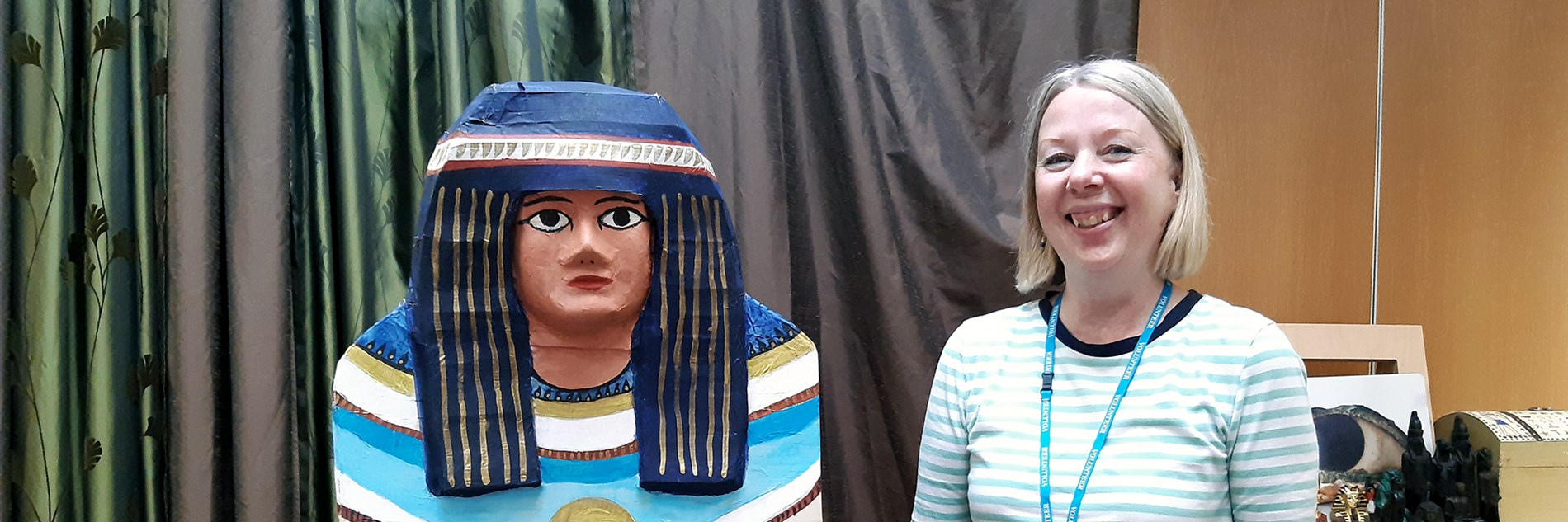 An adult standing next to a replica painted Egyptian coffin, in front of green and white curtain and replica Ancient Egypt artefacts 