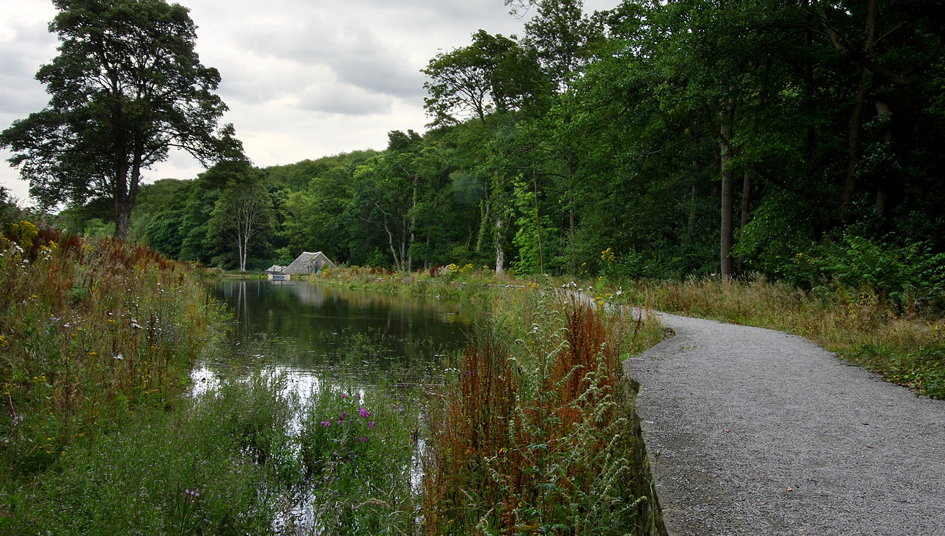 A body of water with a stone path that follows the edge of the water. The path leads to a small building which is surrounded by trees and vegetation. 