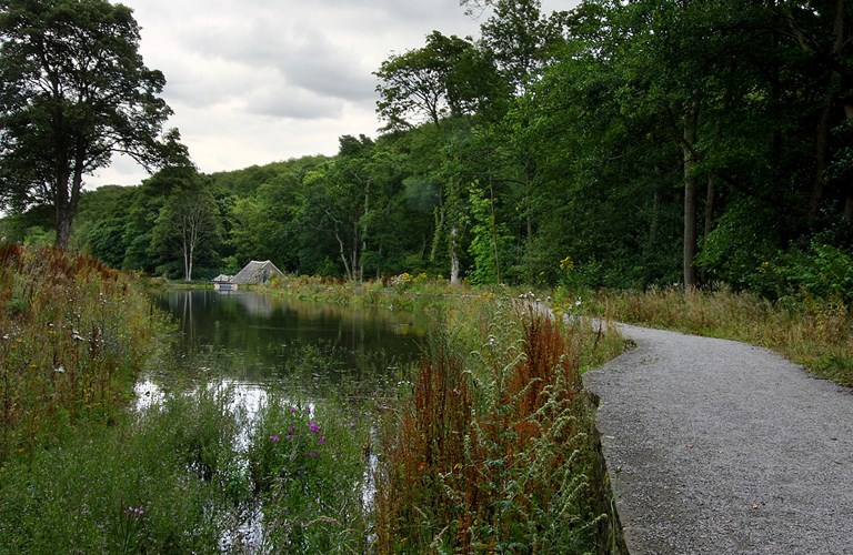 A body of water with a stone path that follows the edge of the water. The path leads to a small building which is surrounded by trees and vegetation. 