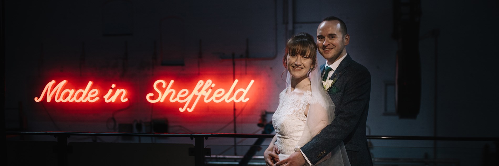 Two adults in wedding outfits in front of a "Made in Sheffield" neon sign inside Kelham Island Museum