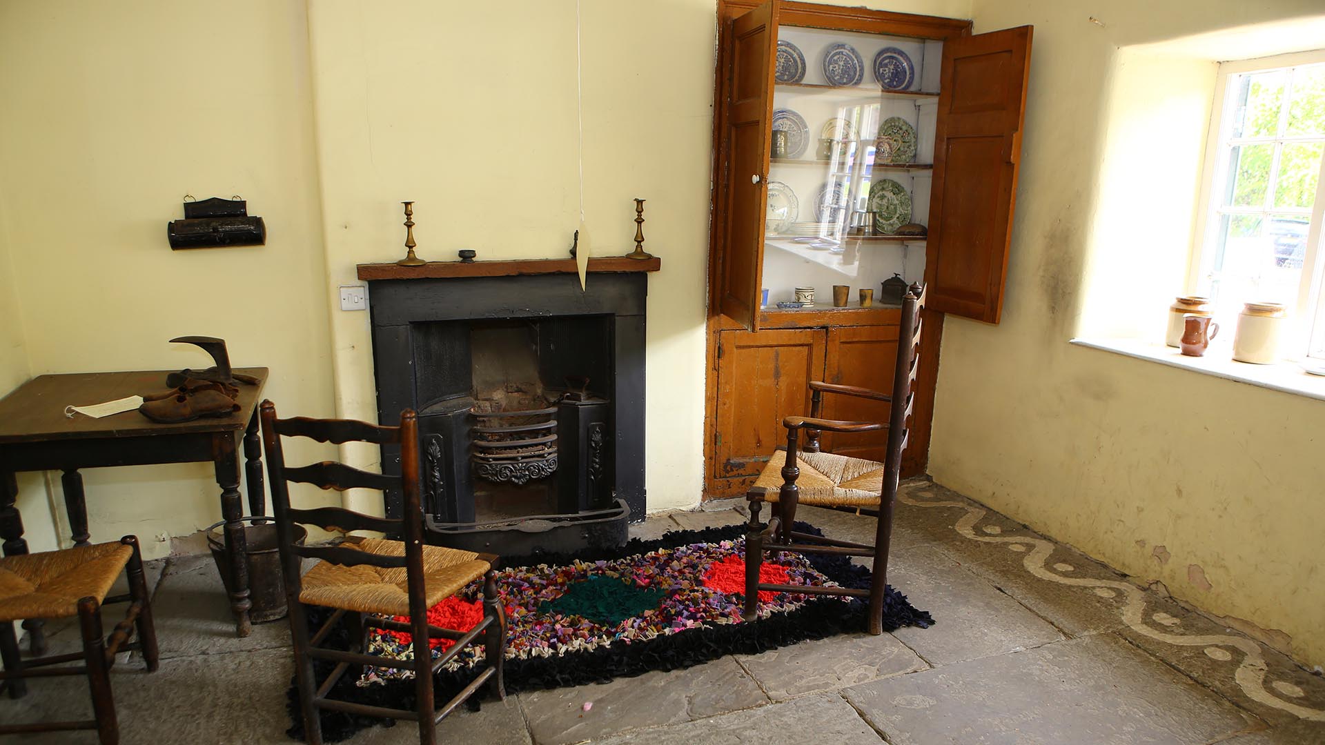 In a room with an open fire grate, two wooden ladderback chairs with woven rush seating stand by a pegged hearthside rug on a stone flagged floor. Alcove cupboards off to one side display antique china crockery. 