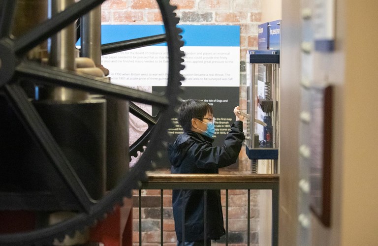 A child wearing a medical facemask operating an interactive scale model of a hydraulic press alongside the real thing.