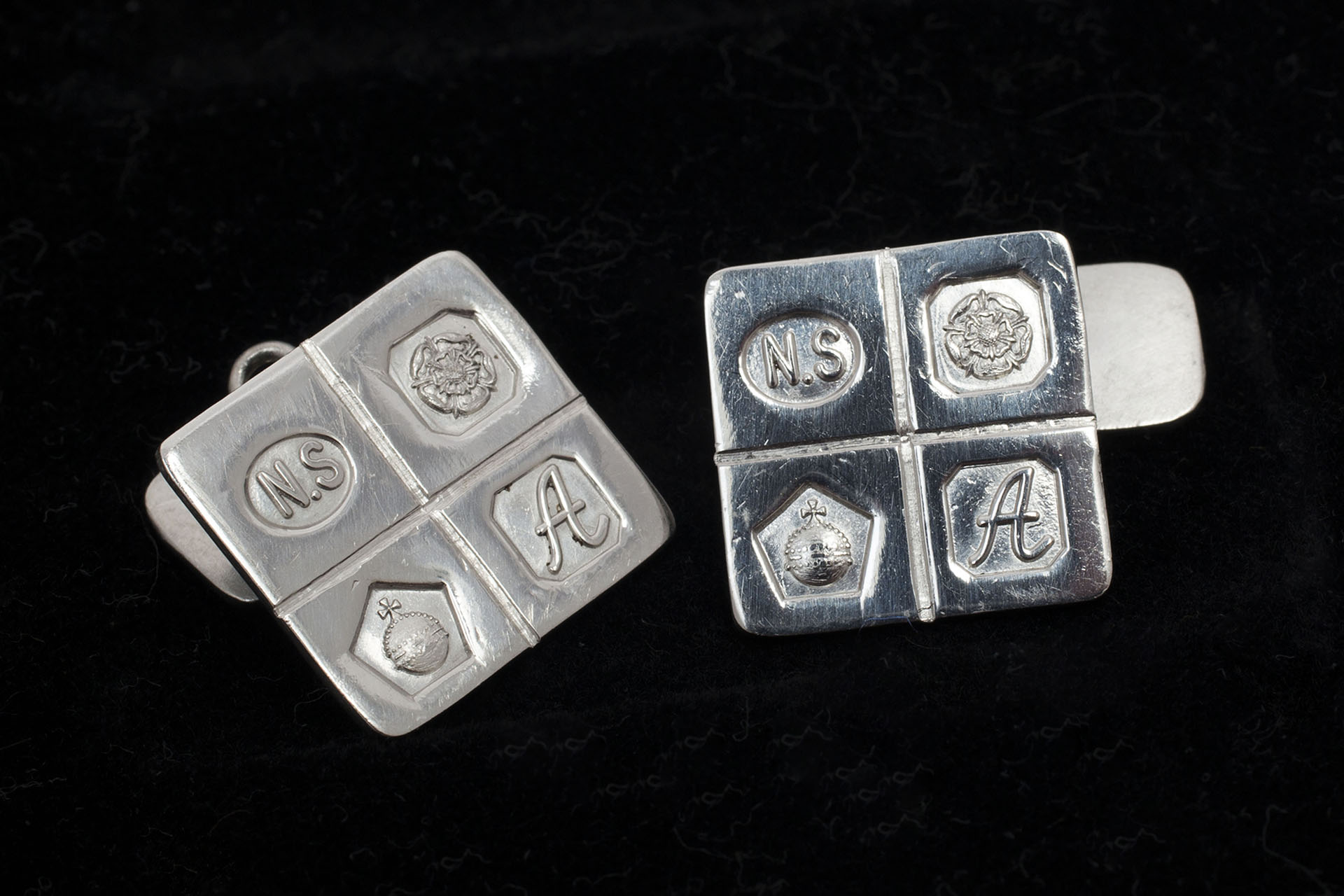 A pair of silver square cufflinks embossed with 4 symbols - the letters N.S, a flower, an orb and the letter A.  