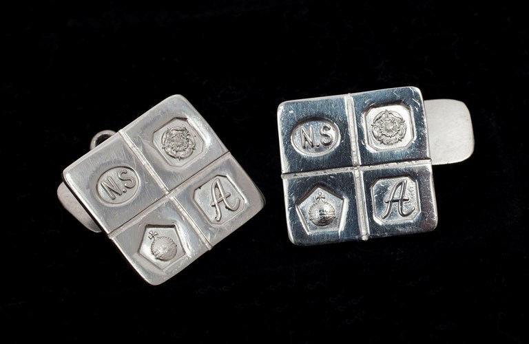 A pair of silver square cufflinks embossed with 4 symbols - the letters N.S, a flower, an orb and the letter A.  