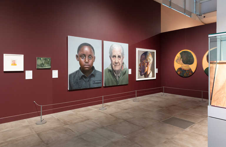 One corner of an exhibition in Millennium Gallery, with ox blood painted walls. There are seven portraits on the walls, varying in size.