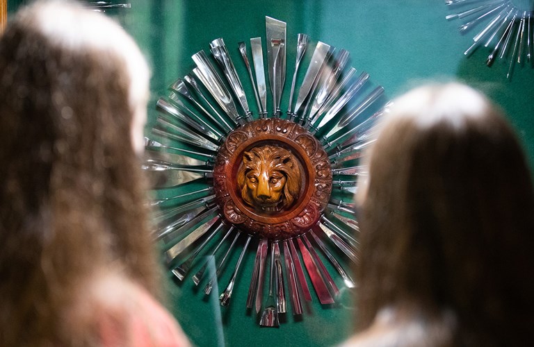 The back of two people's head as they look at a round wooden lion carving with metal tools arranged around it like sun rays.