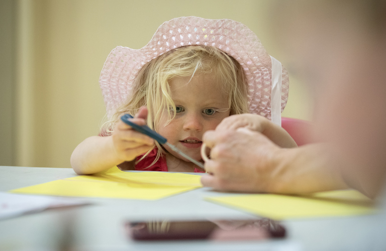 A photograph of a child with blonde hair and a pink straw hat using scissors to cut yellow paper.