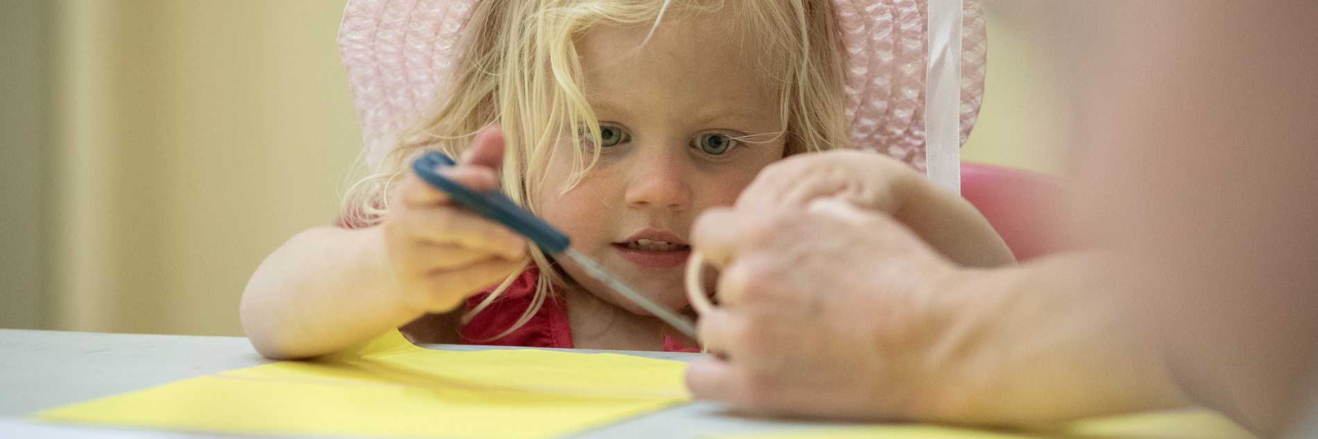 A photograph of a child with blonde hair and a pink straw hat using scissors to cut yellow paper.
