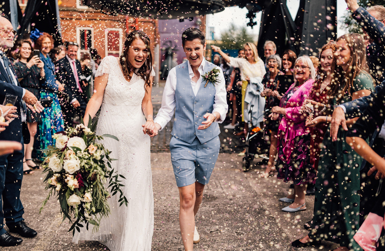 Two adults in wedding outfits being showered with confetti by a crowd of people under the Bessemer Converter