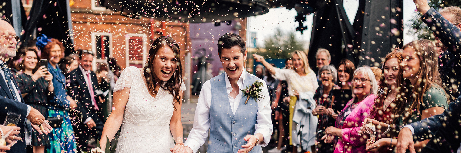 Two adults in wedding outfits being showered with confetti by a crowd of people under the Bessemer Converter