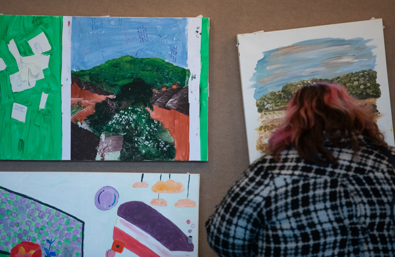 A person with shoulder length pink and black hair and a black and white check shirt in front of a display of three landscape artworks. 
