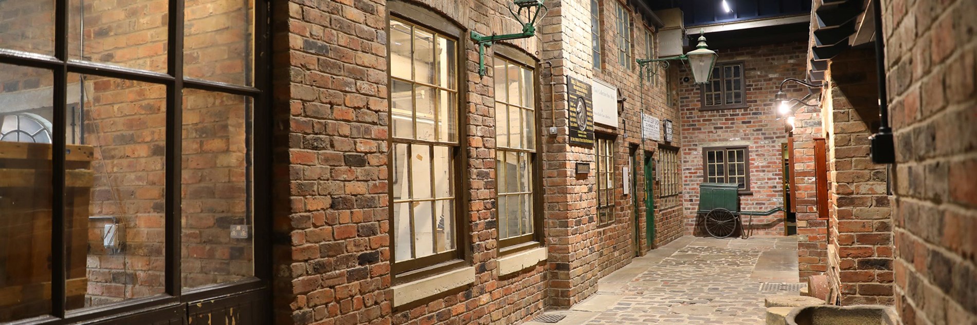 A perspective view along a restored cobbled street. The red brick walled workshops have multi-paned sash windows. There are two green wall mounted gas lamps and a green handcart at the end of the street.