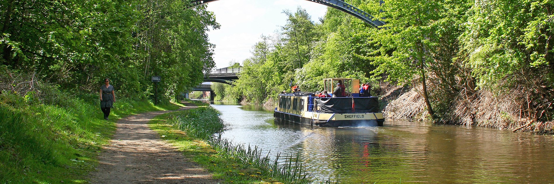 A canal boat on the water going under the first of three bridges on a sunny day. On the left bank a person walks along the footpath which runs next to green trees. On the opposite bank trees and scrub vegetation grow next to the water.
