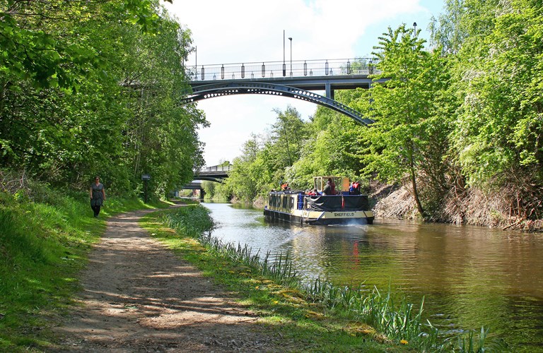 A canal boat on the water going under the first of three bridges on a sunny day. On the left bank a person walks along the footpath which runs next to green trees. On the opposite bank trees and scrub vegetation grow next to the water.