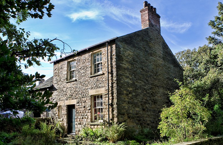 Outside of stone built cottage with Georgian windows and chimney stack in a wooded location in bright sunshine.