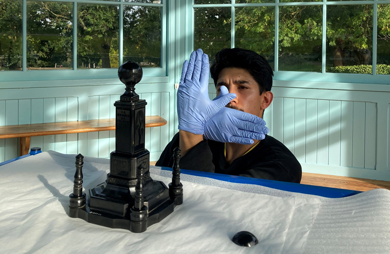 A person behind a table wearing blue gloves making an overlapping hand gesture in an L shape. On the table is a small version of a monument, with four columns surrounding a large central square tiered column topped with a sphere.