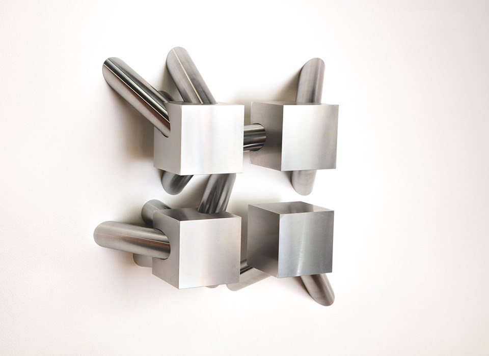 A aluminium metal sculpture of four cubes, arranging in a square shape, with several cylindrical shapes extending from them.