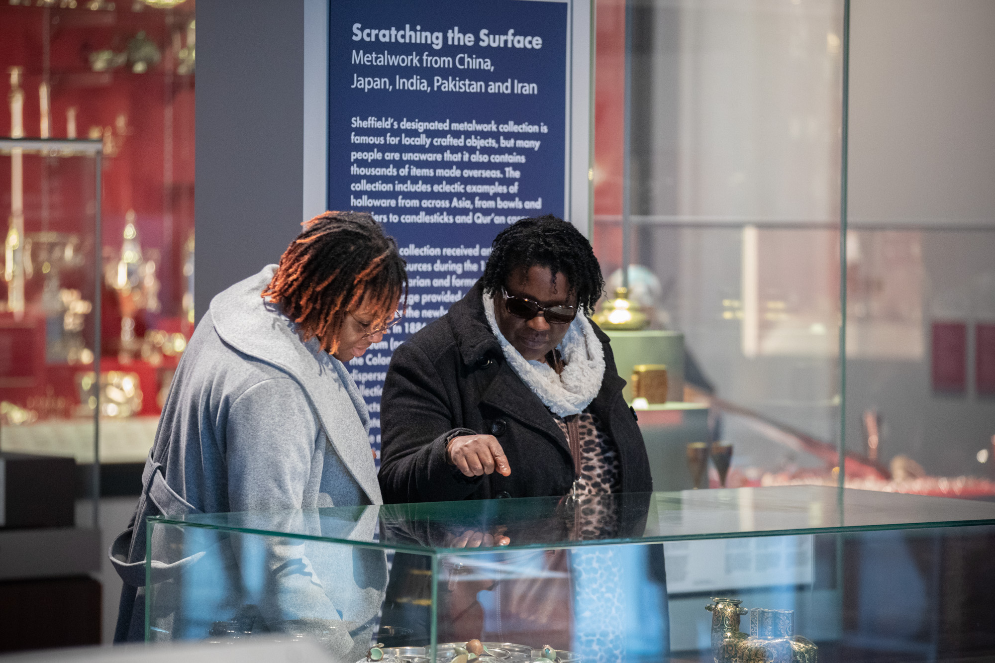 Two adults are looking at a glass cabinet in the gallery, one of the adults is pointing. There is a dark blue gallery sign behind them. 