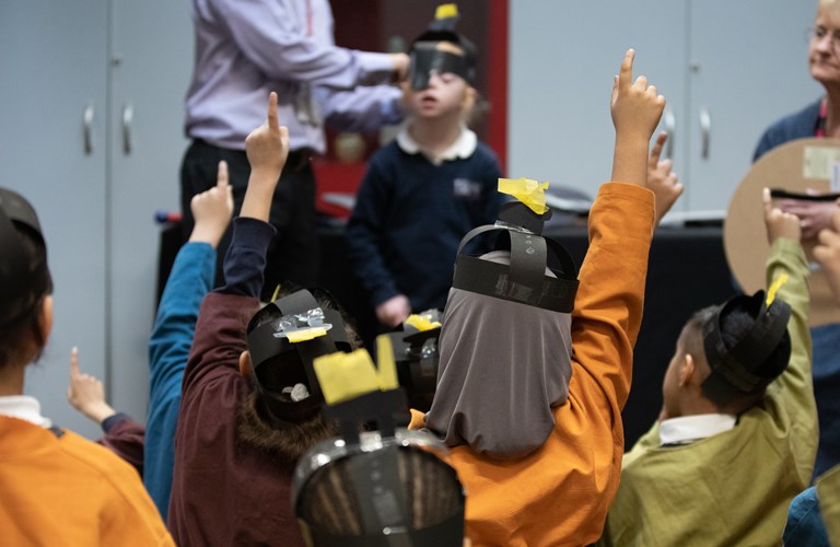 A view from behind of several primary school aged children in a school session, showing their heads, shoulders and raised hands. They are dressed as Anglo Saxons and wearing paper helmets.		
