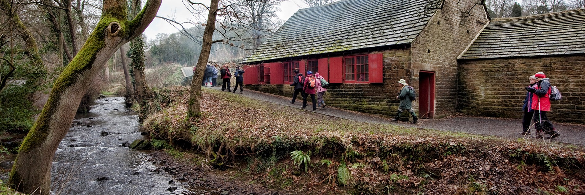 A group of adults walking along a path between a river and a low stone building. There are a number of trees on the banks of the river and a wood in the distance. 