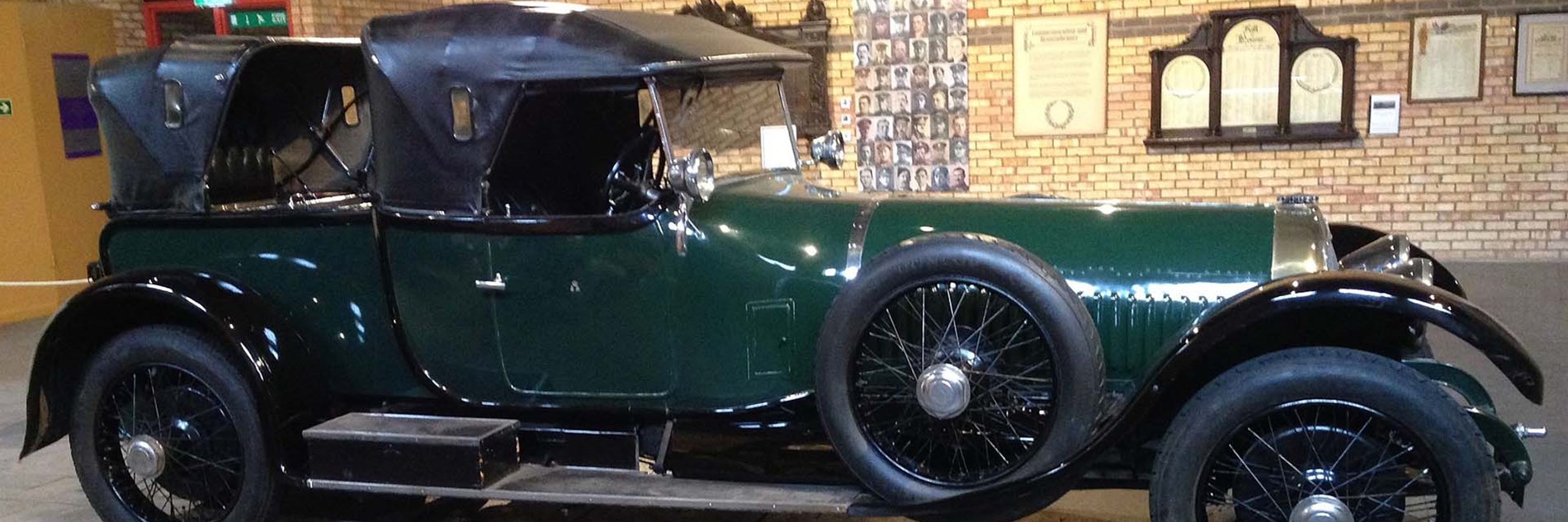 Side elevation of a large vintage car in dark green with a black leather convertible roof, chromed lamps and spoked wheels.