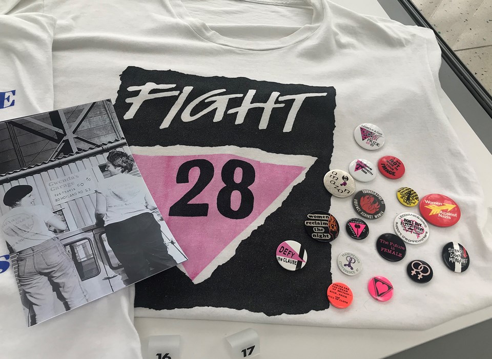 Part of a display in a museum case showing a white t-shirt with a black and pink graphic and the words 'Fight 28'. To the left is a black and white photograph of two female mechanics wearing t-shirts with the Gwenda's Garage logo. To the right is a selection of colourful protest badges.