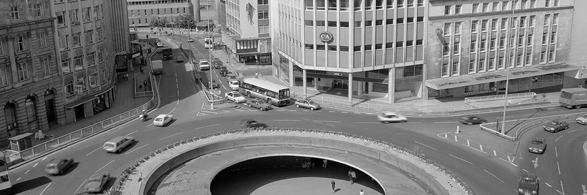 A black and white photograph of a roundabout (known as the “Hole in the Road”) with a view of Sheffield in the background. Cars and buses are driving round the roundabout, and in the middle, below street level, there are people walking and sitting.