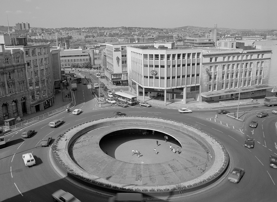 A black and white photograph of a roundabout (known as the “Hole in the Road”) with a view of Sheffield in the background. Cars and buses are driving round the roundabout, and in the middle, below street level, there are people walking and sitting.