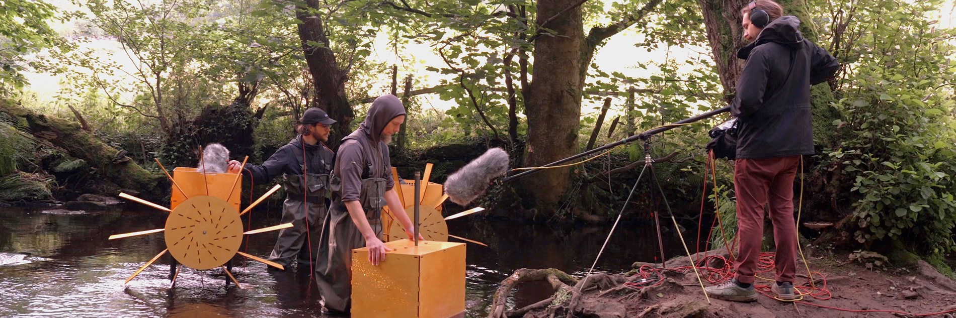 Two musicians playing water-powered mechanical musical instruments on the banks of the River Rivelin. They're wearing waders while someone on the banks of the river records the music using a long-arm microphone over the water.
