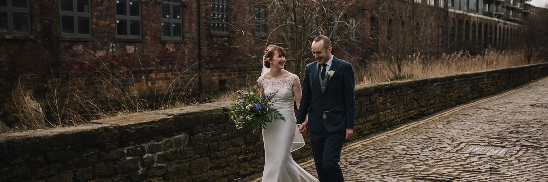 Two adults  in wedding outfits walking on the cobbled road outside Kelham Island Museum