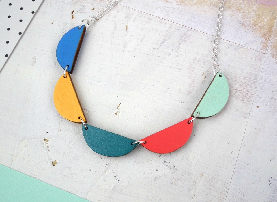A photograph of a necklace consisting of a silver chain and five wooden scallop pieces in different colours. From left to right, the scallops are blue, yellow, turquoise, red and pale blue.