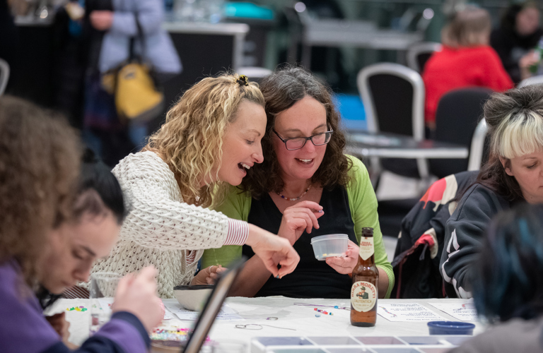 Two women sit next to each other at a table, in conversation. One holds a pot of beads and they are looking through it together. On the table, there are various pots of beads and threads. There is also a bottle of beer.