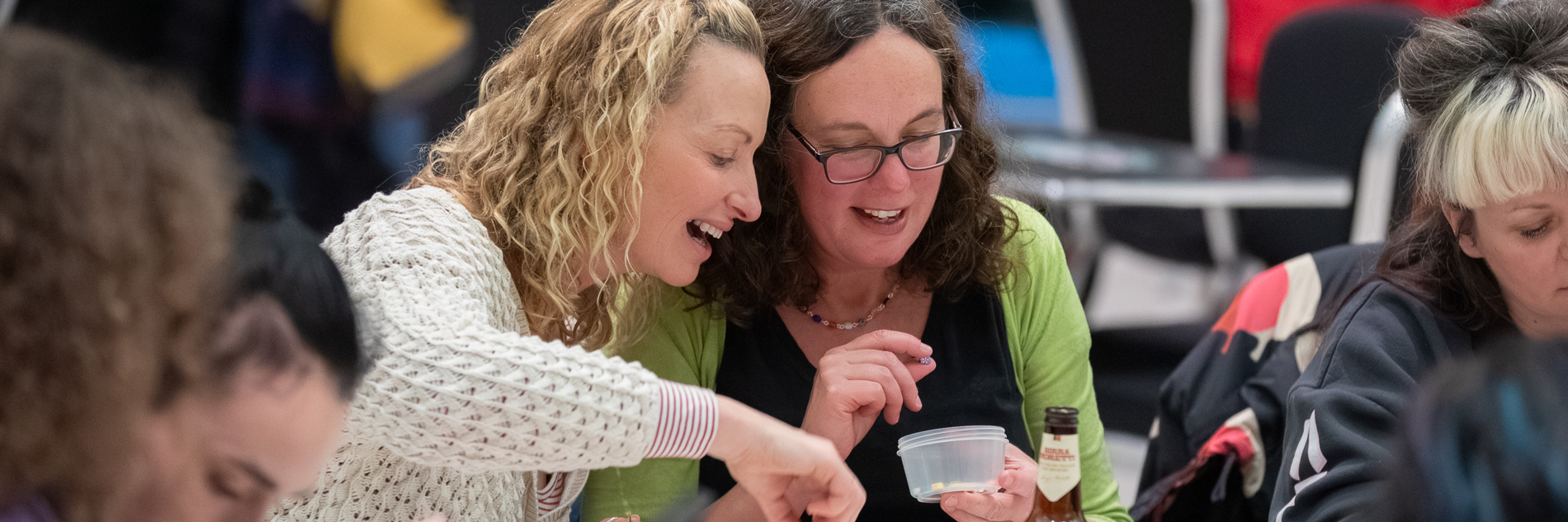 Two women sit next to each other at a table, in conversation. One holds a pot of beads and they are looking through it together. On the table, there are various pots of beads and threads. There is also a bottle of beer.