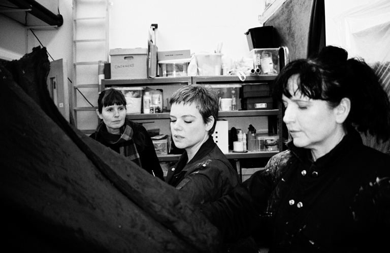 A black and white photograph of three women in a collection storeroom. There are shelves visible behind them, filled with cardboard and plastic boxes. A ladder rests on the wall alongside them.