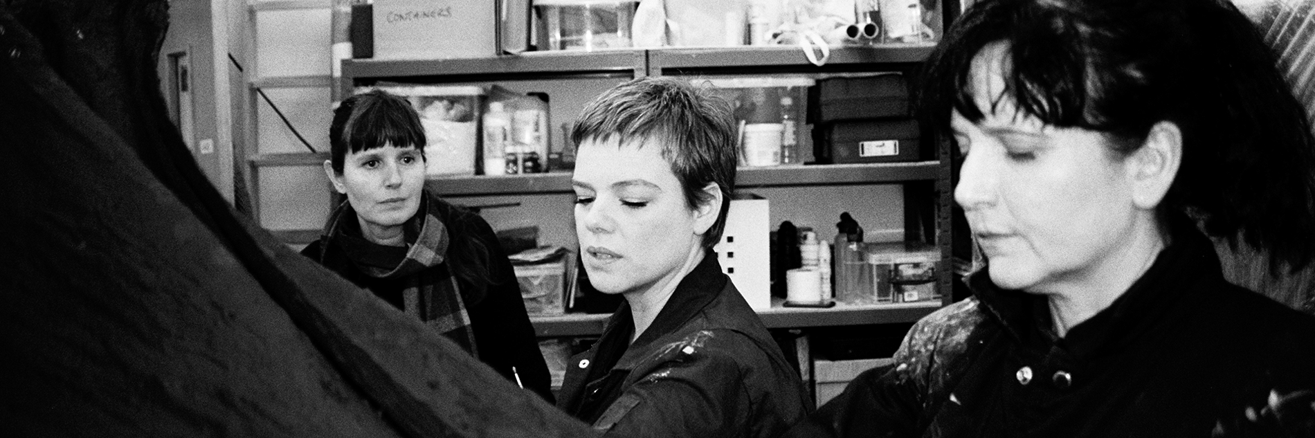 A black and white photograph of three women in a collection storeroom. There are shelves visible behind them, filled with cardboard and plastic boxes. A ladder rests on the wall alongside them.