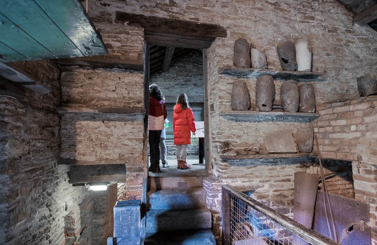 Visitors seen through a doorway with a display of old crucibles on very old wall shelves off to one side.