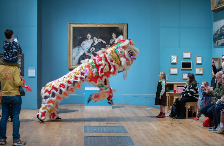 An image of a chinese liondance. The lion is shown jumping into the air with it's front 'legs'. A little girl is stood directly in front of it, looking up with excitement. They are in a picture gallery with blue walls and wooden floor.