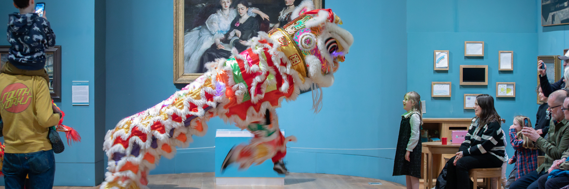 An image of a chinese liondance. The lion is shown jumping into the air with it's front 'legs'. A little girl is stood directly in front of it, looking up with excitement. They are in a picture gallery with blue walls and wooden floor.