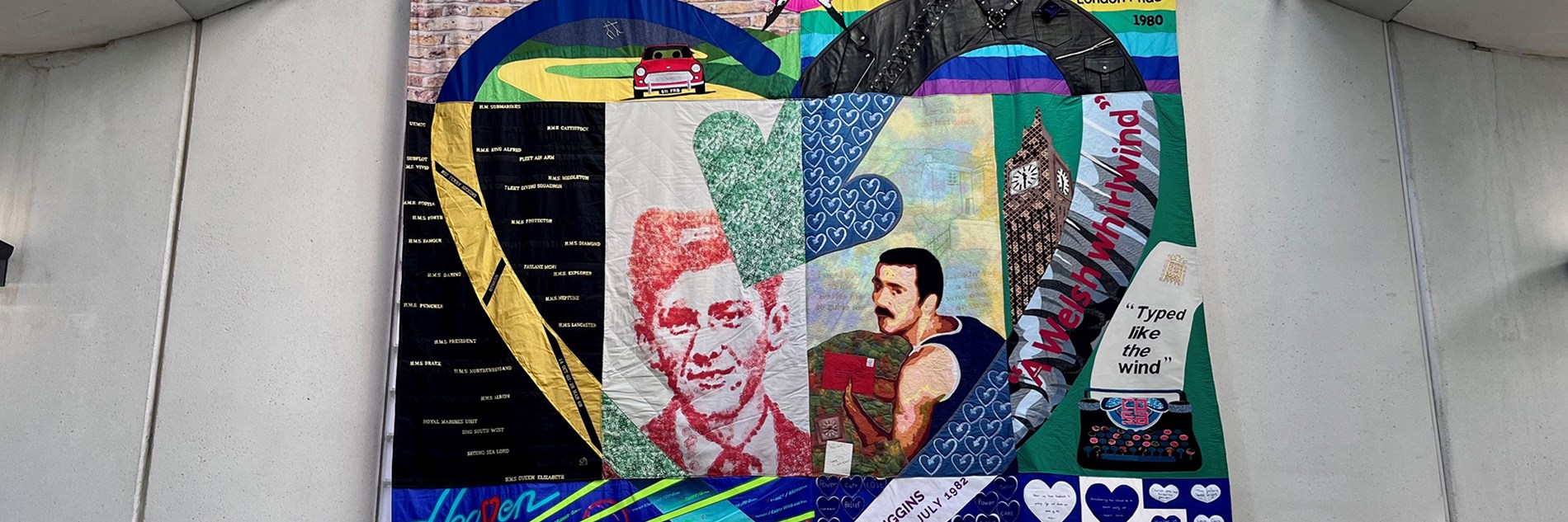 A colourful embroidered quilt featuring text and images. At the centre are two portraits of Terry Higgins which are framed by a heart motif. The quilt is hung on a concrete and glass wall in a contemporary building.