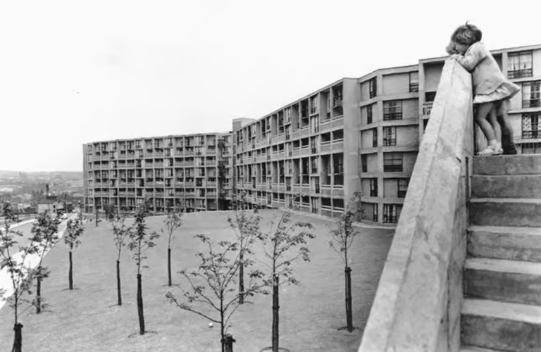 A black and white photograph of Park Hill flats in Sheffield. Trees are visible in the foreground, and a young girl in a dress in visible peeking over a staircase on the right