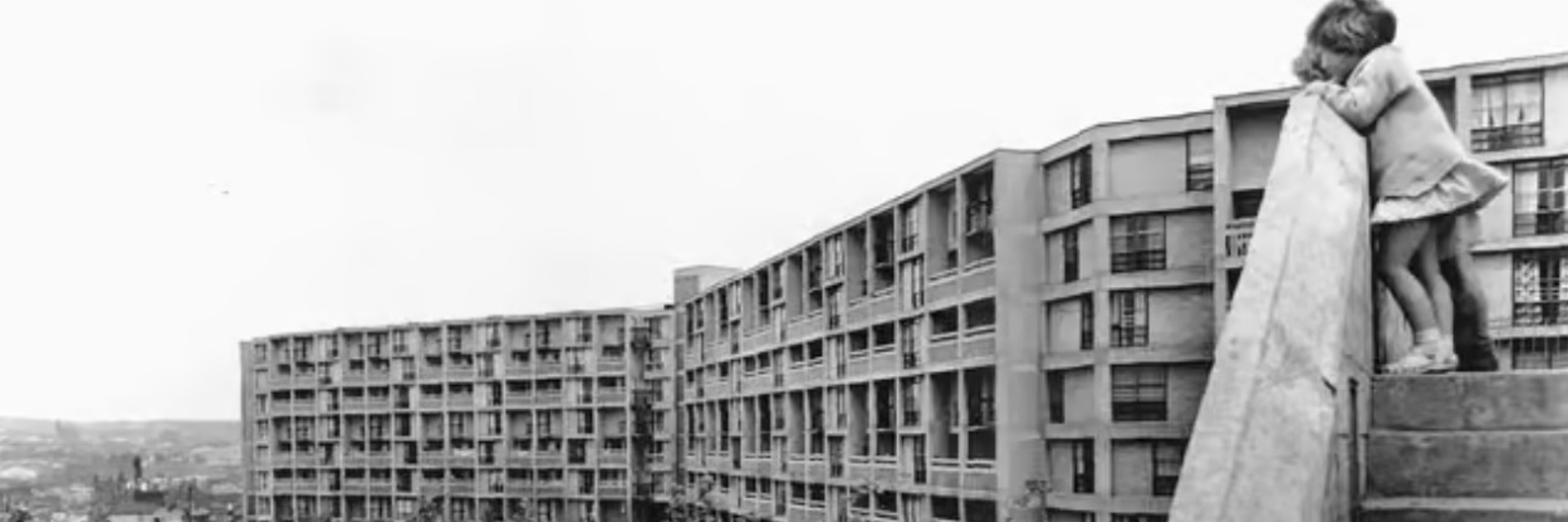 A black and white photograph of Park Hill flats in Sheffield. Trees are visible in the foreground, and a young girl in a dress in visible peeking over a staircase on the right