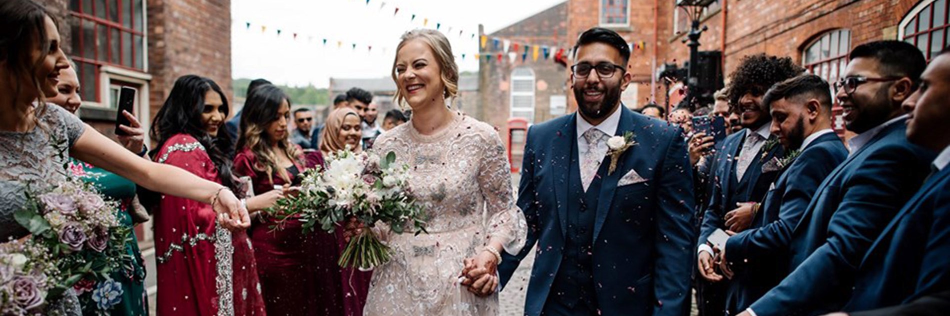 Two adults  in wedding outfits being showered with confetti by a crowd of people outside Kelham Island Museum