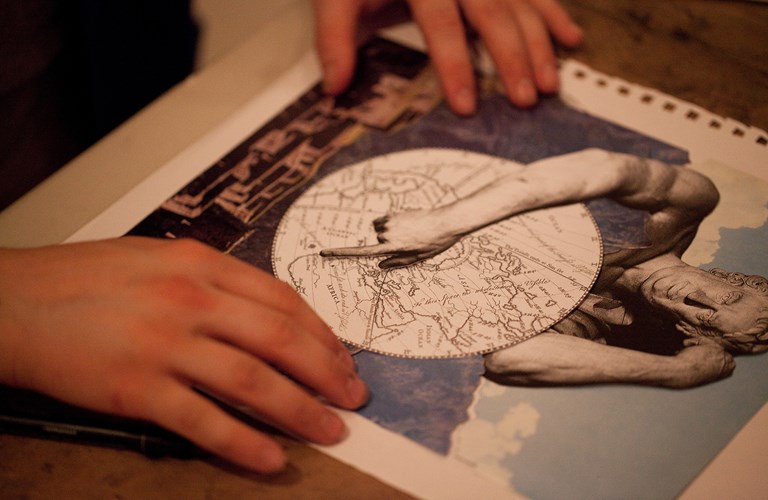 A pair of hands arranging material in a collage, including a map, a photograph of a skyscape and an image of a historic statue.