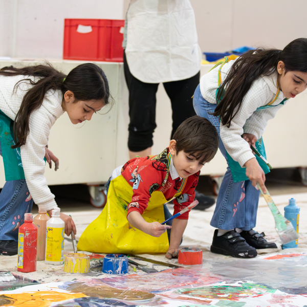 Three children in aprons work on a large collaborative painting on the floor