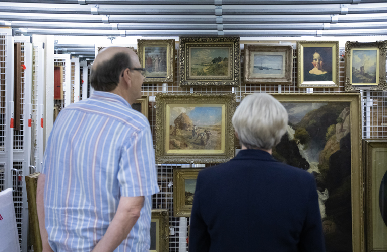 Two adults looking at a collection of framed paintings mounted on a sliding frame