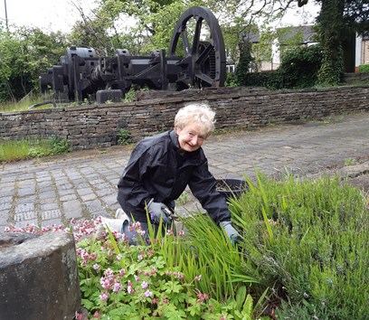 A smiling adult gardening on a cobbled walkway, in front of a cobbled wall. Some historic engineering equipment can be seen in the background 
