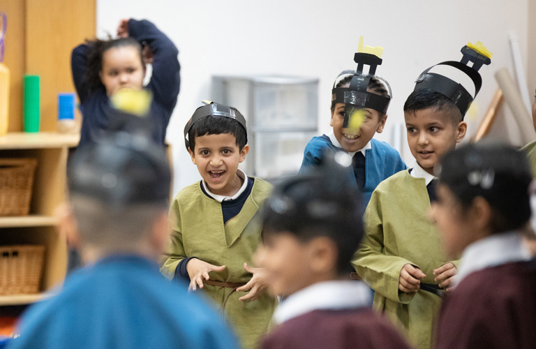 Seven smiling primary school aged children taking part in a school session. They are dressed as Anglo Saxons and wearing brightly coloured tunics and paper helmets which they have made in the session.
