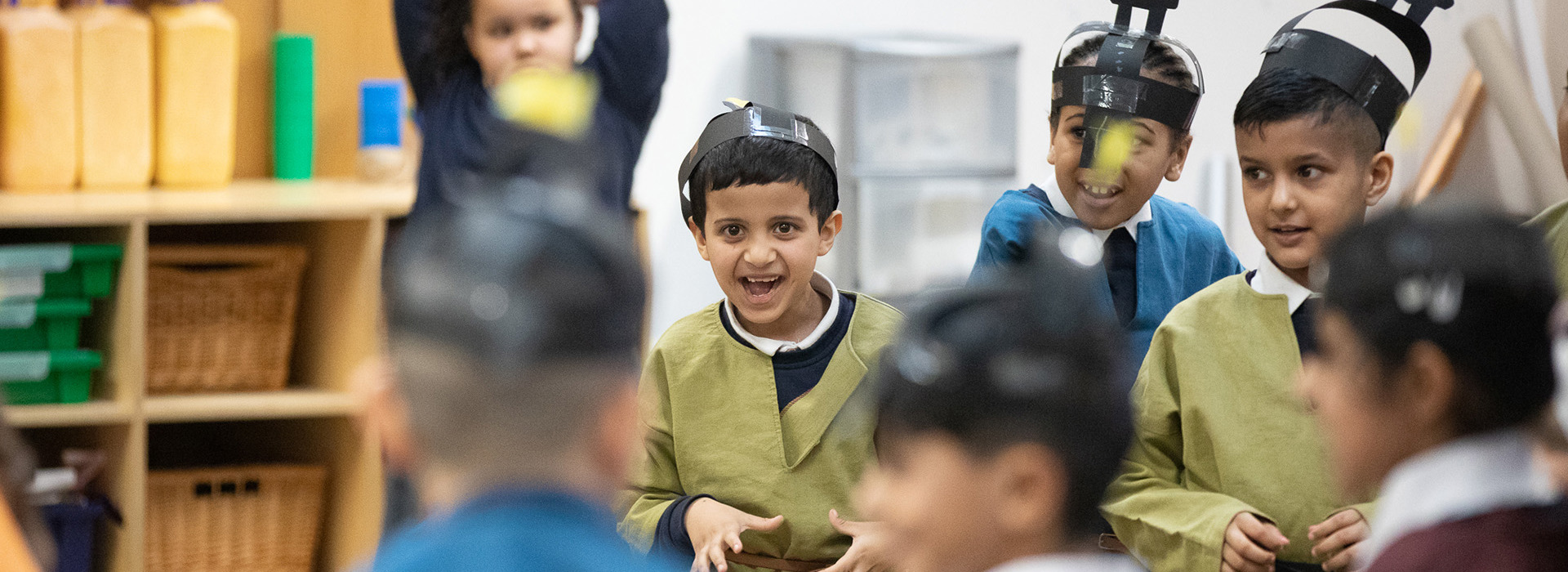 Seven smiling primary school aged children taking part in a school session. They are dressed as Anglo Saxons and wearing brightly coloured tunics and paper helmets which they have made in the session.