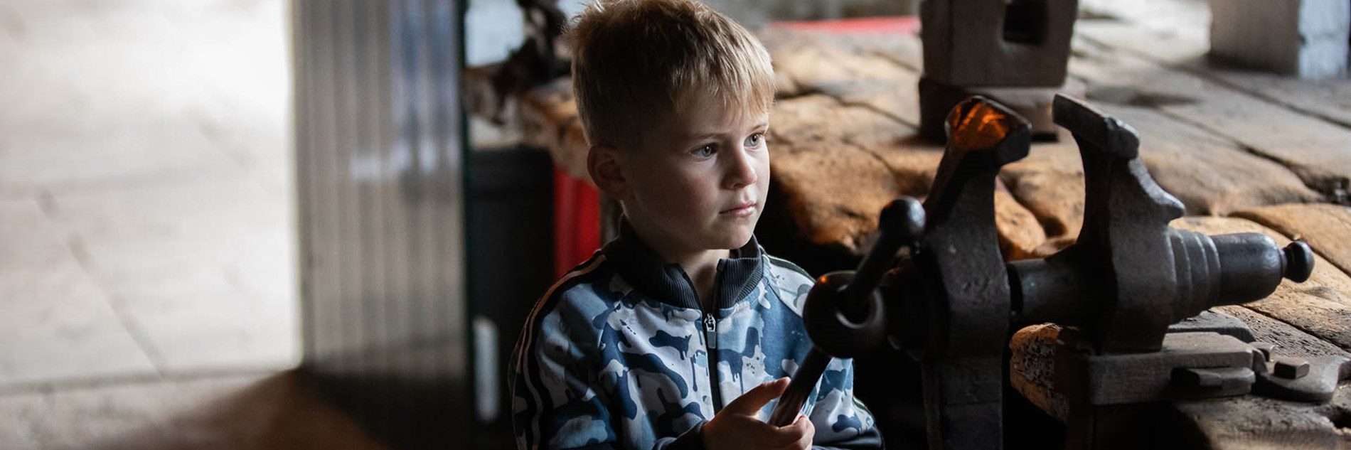 A child visitor looking intently at a large old vice mounted on a very worn workbench.