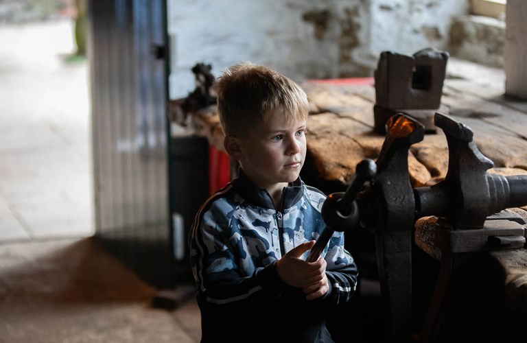 A child visitor looking intently at a large old vice mounted on a very worn workbench.